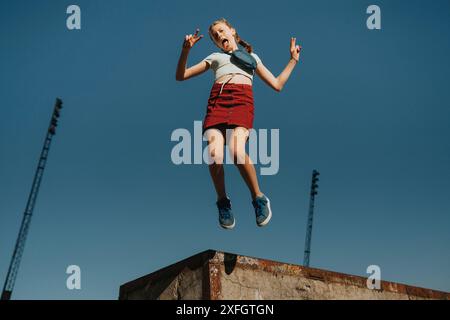 Sorgloses Mädchen, das mitten in der Luft springt, während es ein Friedenszeichen gegen den blauen Himmel macht Stockfoto