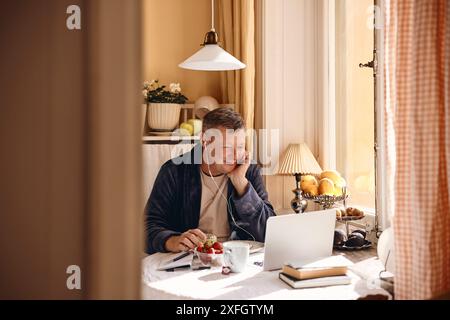 Lächelnder Mann mit kabelgebundenen in-Ear-Kopfhörern und beobachtete den Laptop, während er am Esstisch neben dem Fenster saß Stockfoto