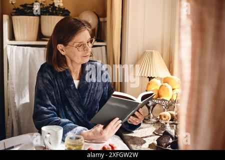 Eine ältere Frau liest Buch, während sie zu Hause am Esstisch sitzt Stockfoto