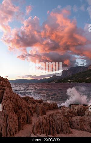 Farbenfroher Sonnenuntergang auf dem Meer. Rot-orange Wolken, Wellen brechen gegen die Felsen, gefärbt von der untergehenden Sonne. Die Küste Kroatiens. Stockfoto