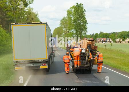 Tarnow, Polen - 19. Mai 2023: Straßenarbeiter in reflektierenden Westen bedienen auf einer Landstraße Straßenpflastermaschinen mit vorbeifahrenden Fahrzeugen. Stockfoto