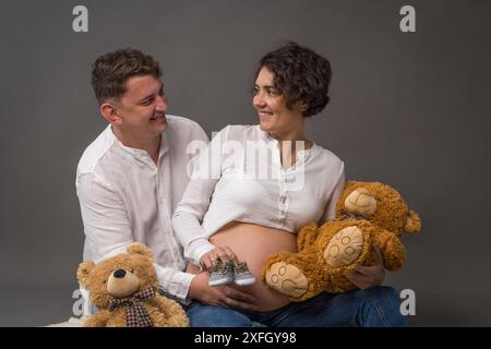 Eine glückliche schwangere Frau und ihr Mann sitzen auf dem Boden und halten zwei Teddybären in der Hand. Sie sehen sich mit Liebe an. Husban Stockfoto