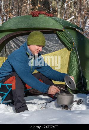 Ein Mann mittleren Alters kocht Essen auf dem Herd im Winterwald. Titantopf. Warten auf Essen. Frühstück im Zelt Stockfoto