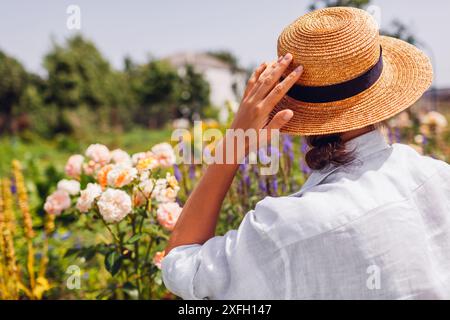 Rückansicht einer Gärtnerin mit Strohhut, die im Sommergarten Rosenblumen genießt. Bewundern Sie die Landschaft Stockfoto