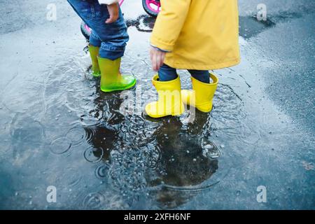 Zwei Kinder in Regenstiefeln stehen auf nassem Asphalt. Eine in gelber Kleidung spritzt mit dem linken Fuß, die andere trägt grüne Stiefel und blaue Jeans Stockfoto
