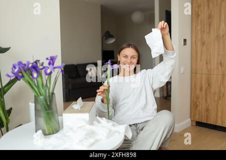 Allergiefrei. Eine fröhliche junge Frau wirft zu Hause Servietten hoch. Glückliches Leben ohne laufende Nase, Juckreiz oder Husten Symptome. Allergenstrauß von Irisblumen in der Vase, benutzte Taschentücher auf dem Tisch. Stockfoto