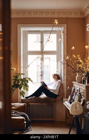 Seitenansicht einer älteren Frau, die Buch liest, während sie auf der Fensterbank sitzt Stockfoto