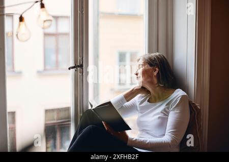 Eine ältere Frau, die durch das Fenster schaut, während sie zu Hause ein Buch hält Stockfoto
