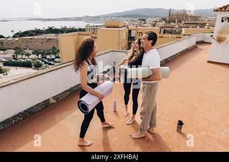 Glücklicher Mann, der eine Trainingsmatte hält und mit weiblichen Freunden nach dem Yoga-Kurs auf dem Dach steht Stockfoto