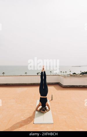 Junge Frau, die Kopfstand übt, während sie Yoga auf dem Dach am Himmel macht Stockfoto