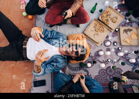Direkt über dem Schuss eines glücklichen Mannes, der auf einer Decke liegt, von Freunden auf dem Dach Stockfoto
