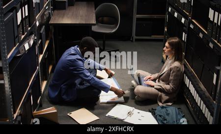 Ein afroamerikanischer Polizist liest Akten im Büro und versucht, Details und Hinweise aufzudecken. Multiethnische Detektive studieren Fallinformationen, suchen nach forensischen Beweisen. Stockfoto