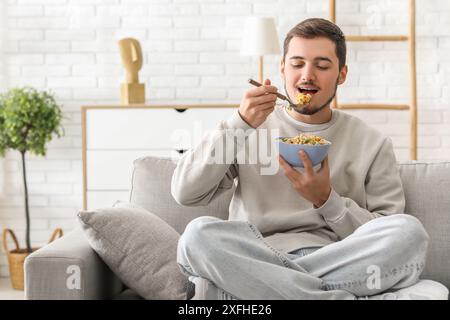 Hübscher junger Mann, der leckere Müsliringe isst, während er auf dem Sofa im Wohnzimmer sitzt Stockfoto