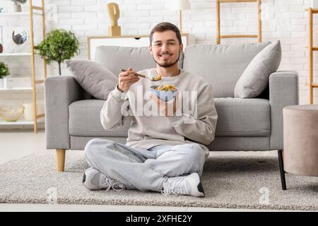 Hübscher junger Mann, der leckere Müsliringe isst, während er auf dem Boden im Wohnzimmer sitzt Stockfoto