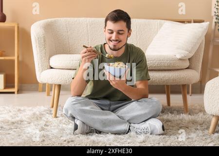 Hübscher junger Mann, der leckere Müsliringe isst, während er auf dem Boden im Wohnzimmer sitzt Stockfoto
