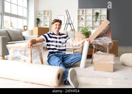 Junger Mann mit einer Rolle Stretchfolie und eingewickelten Möbeln, der am Umzugstag im Zimmer sitzt Stockfoto