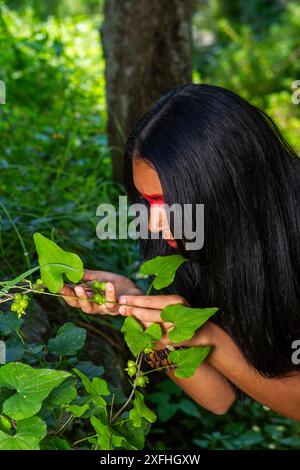 Eine junge indigene Frau mit bemaltem Gesicht inspiziert sorgfältig grüne Beeren in einer üppigen Waldlandschaft. Stockfoto