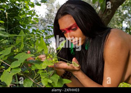 Eine junge indigene Frau aus einem brasilianischen Stamm mit gemaltem Gesicht untersucht sorgfältig grüne Beeren in einer üppigen Waldlandschaft. Stockfoto