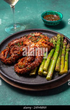 Gegrillter Tintenfisch mit Spargel, serviert auf einem schwarzen Teller, der lebhafte Farben und Texturen vor türkisfarbenem Hintergrund hervorhebt. Stockfoto