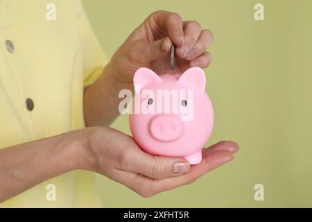 Frau, die Münze in rosa Schweinebank auf olivfarbenem Hintergrund steckt, Nahaufnahme Stockfoto