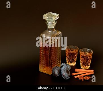 Pflaumenlikör mit Zimt und Anis, hausgemachter Alkohol in einer Kristallkaraffe und zwei Gläser auf dunklem Hintergrund, neben Pflaumenfrüchten und Gewürzen. Clo Stockfoto