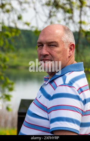 Ein kahlköpfiger Mann mittleren Alters in einem gestreiften Hemd steht in der Nähe eines Sees. Stockfoto