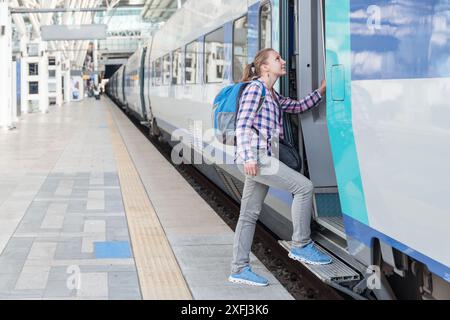 Junge weibliche Touristen, die in den Zug einsteigen. Lächelnde hübsche Frau mit blauem Rucksack am Bahnhof. Bahnfahrt. Moderner Hochgeschwindigkeitszug. Stockfoto