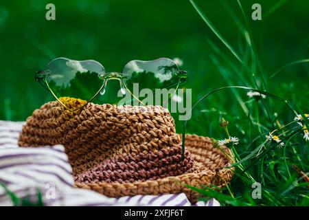 Strohhut im Boho-Stil und eine grüne Sonnenbrille in Herzform liegen auf einem grünen Rasen mit blühender Wiese. Sommerreise, Reise. Sommer Natur. Stockfoto