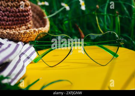 Strohhut im Boho-Stil und eine grüne Sonnenbrille in Herzform liegen auf einem grünen Rasen mit blühender Wiese. Sommerreise, Reise. Sommer Natur. Stockfoto