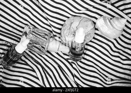 Serumflaschen aus Glas ruhen auf gestreiftem schwarz-weißem Stoff von oben. Sommerliche Tagespflege, weibliche Routine. Kosmetikprodukte, Muschelflachlage. Stockfoto