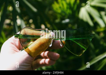 Eine Frau, die an sonnigen Sommertagen durch eine grüne, herzförmige Sonnenbrille auf die Natur schaut. Urlaub am Meer im tropischen Resort. Exotische Landschaft. Stockfoto