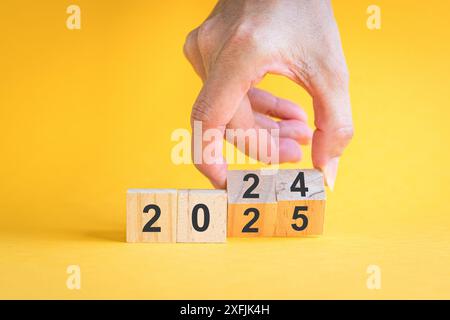 Finger drehen sich von 2024 auf 2025, Zahlen auf Holzblöcken. Auf gelbem Hintergrund, Neujahrskonzept. Stockfoto
