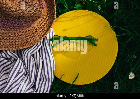 Strohhut im Boho-Stil und eine grüne Sonnenbrille in Herzform liegen auf einem grünen Rasen mit blühender Wiese. Sommerreise, Reise. Sommer Natur. Stockfoto