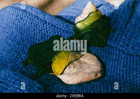 Grüne Sonnenbrille in Herzform, Muschel auf blauem Textil. Urlaub am Meer, Meer im Sommer. Entspannen im Sommer. Weibliches Zubehör. Sonniger Tag. Stockfoto