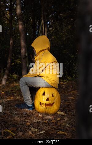 Das Kind in orangefarbener Jacke und Kapuze sitzt auf einem großen Kürbis mit geschnitzter Grimasse. Abendwald, Kind durch Lichtstreifen hervorgehoben. Halloween-Feiertage. mo Stockfoto