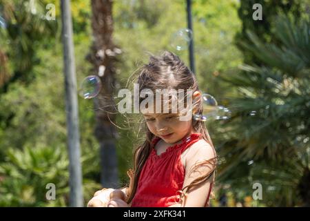 Das kleine Mädchen in leuchtendem rotem Kleid hält ihre Puppe in einem sonnigen Park. Umgeben von Seifenblasen schaffen das grüne Gras und die hohen Palmen ein magisches und Stockfoto