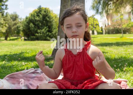 Schönes kleines Mädchen in rotem Kleid in Meditationshaltung, in Nahaufnahme an einem warmen Tag gefangen. Umgeben von grünem Gras vermittelt sie Ruhe und Ruhe Stockfoto