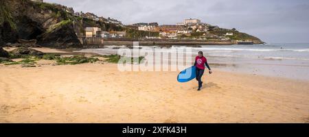 Ein Panoramabild einer Surferin, die ihr Surfbrett nach dem Surfen am Towan Beach in Newquay in Cornwall in Großbritannien trägt. Stockfoto