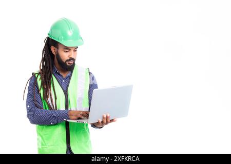 Karibischer Mann mit Dreadlocks und Bart, verheirateter Architekt, der mit Laptop arbeitet, trägt ein blaues Hemd, eine phosphoreszierende grüne Weste und einen grünen Helm Stockfoto