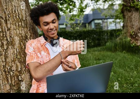 Junger Mann, Afroamerikaner, sitzt unter Baum und benutzt Laptop im Park. Stockfoto