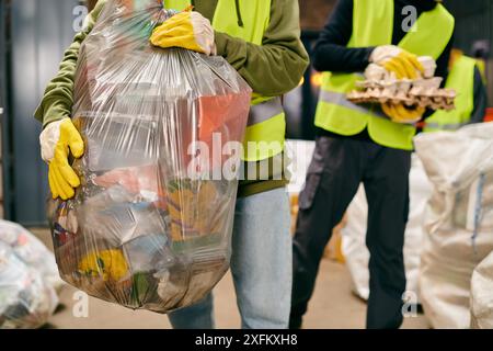 Zwei junge Freiwillige in Sicherheitswesten, Handschuhen und einer riesigen Tüte Müll beim Sortieren von Müll. Stockfoto