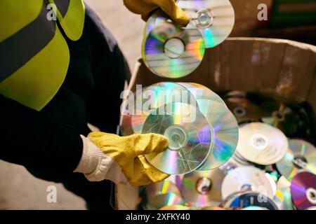 Ein junger Freiwilliger in Handschuhen und Sicherheitsweste, der einen Haufen CDs hält und Abfall im Rahmen umweltbewusster Bemühungen sortiert. Stockfoto