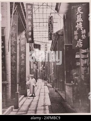 Vintage-Foto der Physic Street (Tsiang-Lan-Kiai) im Kanton (Guangzhou). Illustrationen von Qing China, 1875 ... die Straßen einer chinesischen Stadt unterscheiden sich stark von denen Europas und sind immer extrem eng, außer in Nankin und Peking. Sie sind kreuzweise mit Steinplatten gepflastert, die normalerweise vom Verkehr bis zu einer Höhle in der Mitte des Weges abgetragen werden, und dieser unangenehme Ersatz für die Regenrinnen europäischer Durchgangsstraßen ist das einzige Mittel, mit dem das Regenwasser abtransportiert wird. Die Geschäfte in guten Straßen sind fast einheitlich groß; jedes Gebäude wird von einer Backsteinmauer getrennt Stockfoto