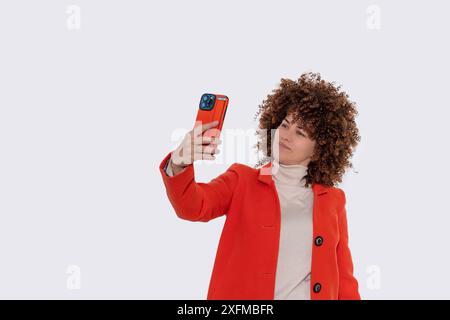 Positive, süße Frau im orangefarbenen Mantel, die ein Handy hält, plaudert, Selfie auf einem Studio-Porträt mit weißem Hintergrund macht Stockfoto