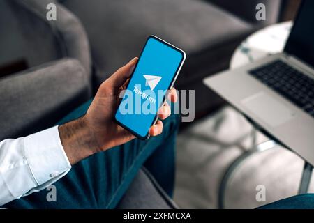 Mann mit einem Handy, das die Telegramm-App auf dem Bildschirm anzeigt. Mann am Arbeitsplatz mit Laptop und Smartphone in der Hand. ROSARIO, ARGENTINIEN - 03. JULI 2024. Stockfoto