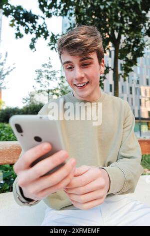 Vertikaler Teenager, der Spaß hat, draußen ein Handy zu benutzen. Sorgloser Kerl lächelt und surft im Internet mit einer Handy-App. Ein echter junger Mann, der zusieht Stockfoto