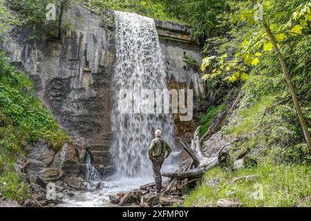 Abseits der ausgetretenen Pfade steht ein alter, erfahrener Wanderer in einer Schlucht unter einem Wasserfall und schaut auf das kaskadierende Wasser. Stockfoto