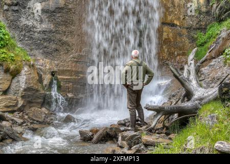 Abseits der ausgetretenen Pfade steht ein alter, erfahrener Wanderer in einer Schlucht unter einem tosenden Wasserfall. Stockfoto