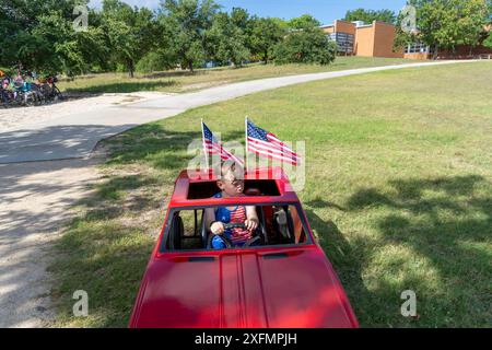 Austin, Texas, USA, 4. Juli 2024. Während der jährlichen Parade am 4. Juli in Barton Hills sitzt ein Junge in einem Spielzeugauto mit fliegenden amerikanischen Fahnen auf dem Spielplatz der Barton Hills Elementary School. Ein paar hundert Einwohner gingen auf der 1/2 km langen Paradestraße zum Schulgelände, wo sie Kekse, Wassermelonen und Eis genossen. Quelle: Bob Daemmrich/Alamy Live News Stockfoto