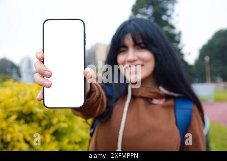 Unscharfe glückliche Frau, die im Park lächelt und ein Telefon mit leerem Bildschirm hält, schaut in die Kamera, die ihr Telefon, ihr Technologiekonzept und ihren Kopierraum zeigt. Stockfoto